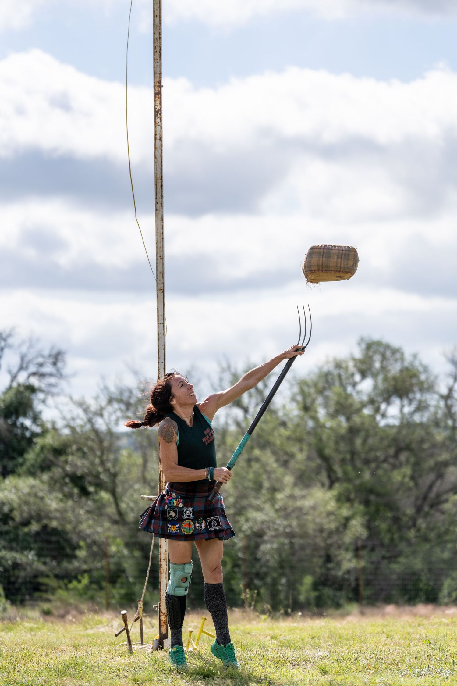 Woman in kilt tossing a sheaf with a pitchfork at a Highland Games competition outdoors.
