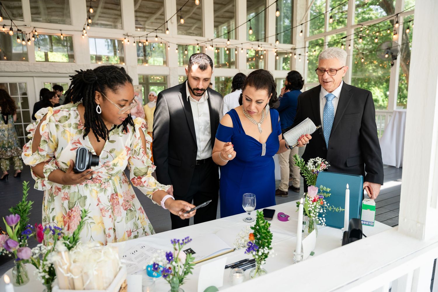 Guests in formal attire gather around a floral-decorated table at an elegant wedding reception venue.