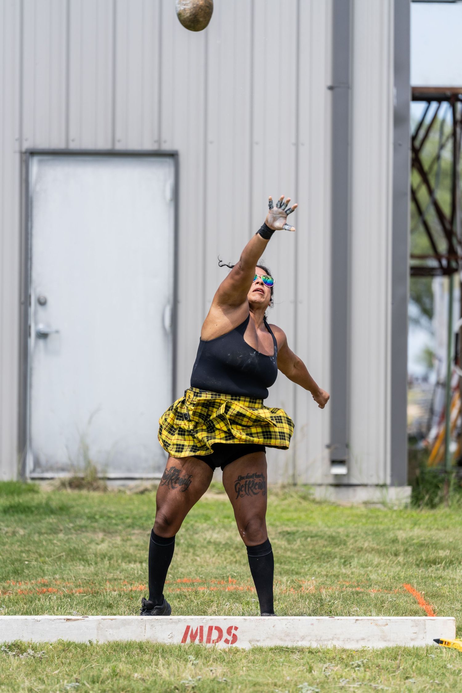 Woman in plaid skirt throwing a heavy ball at a Highland Games athletic competition outdoors.