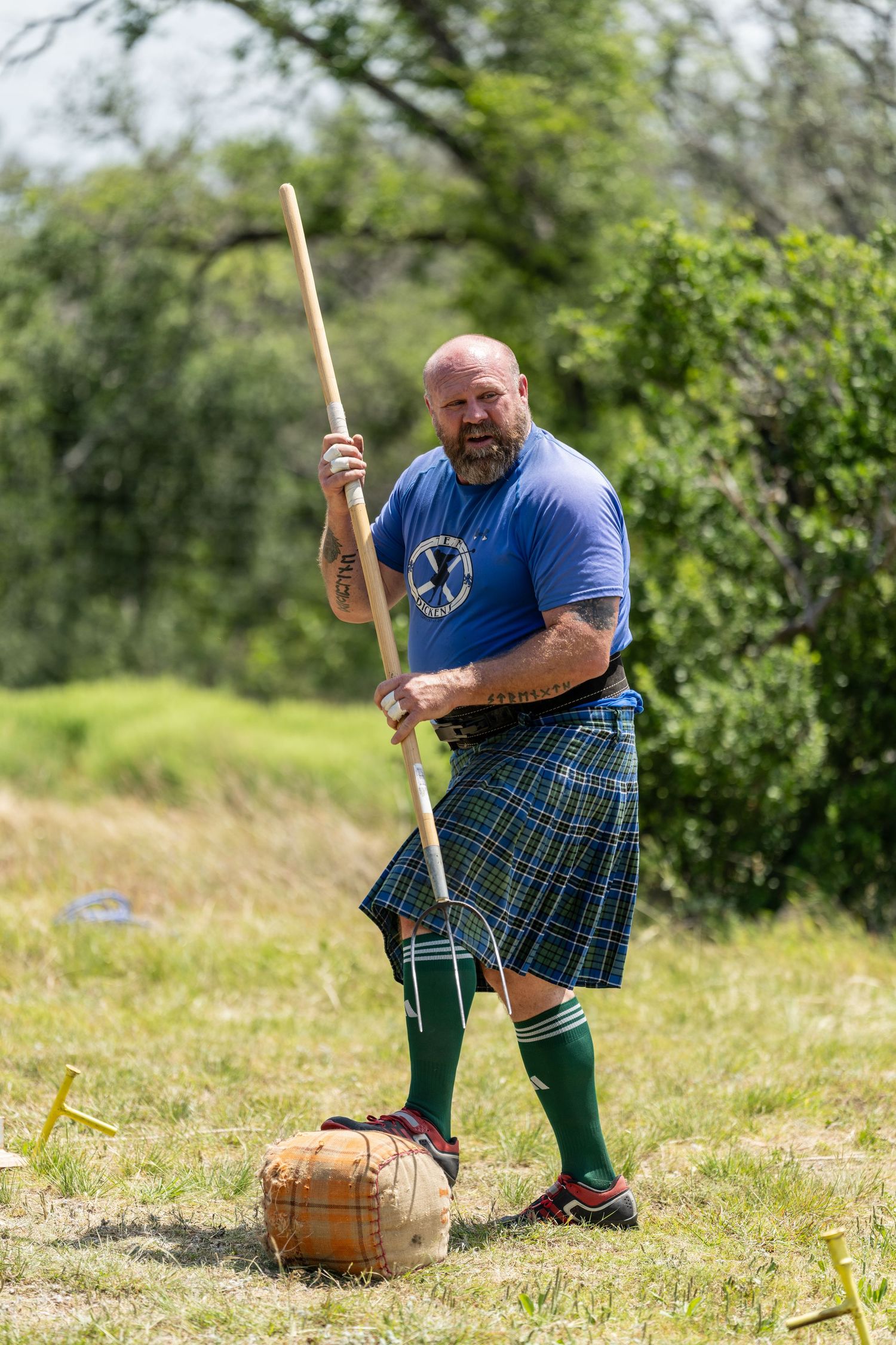 Man in blue shirt and tartan kilt competes in Highland Games, holding a wooden pole beside a weighted bag.