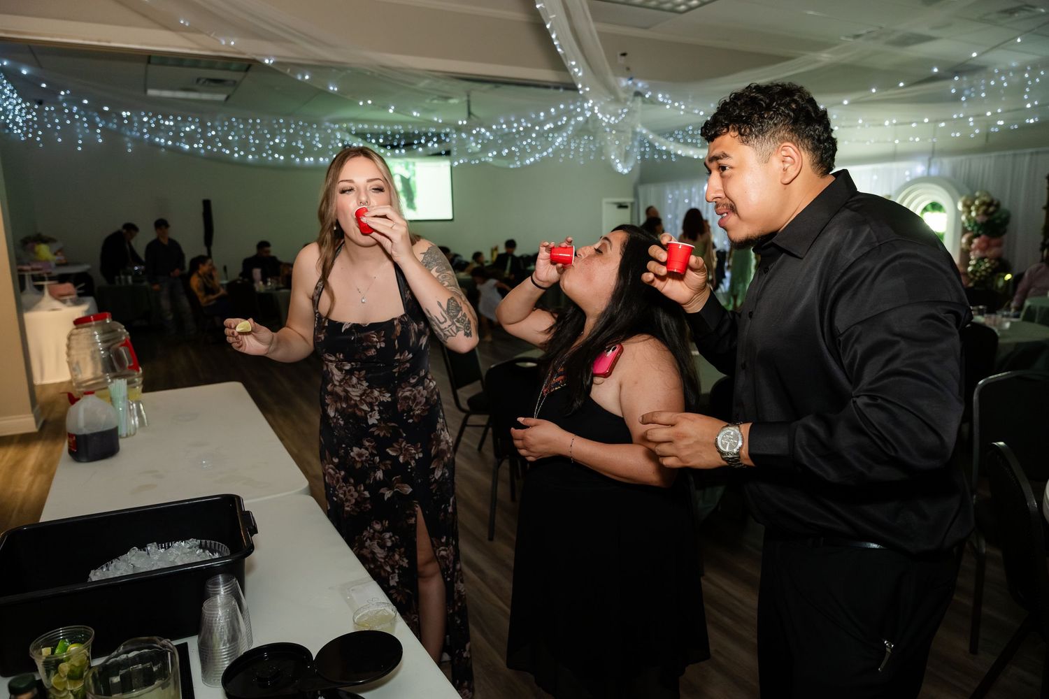 Three guests drinking shots at a party bar in a decorated event venue with fairy lights.