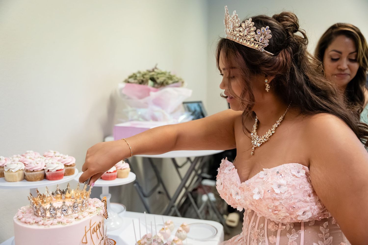 Young woman in pink gown and tiara cuts cake at quinceañera celebration with cupcakes on display.