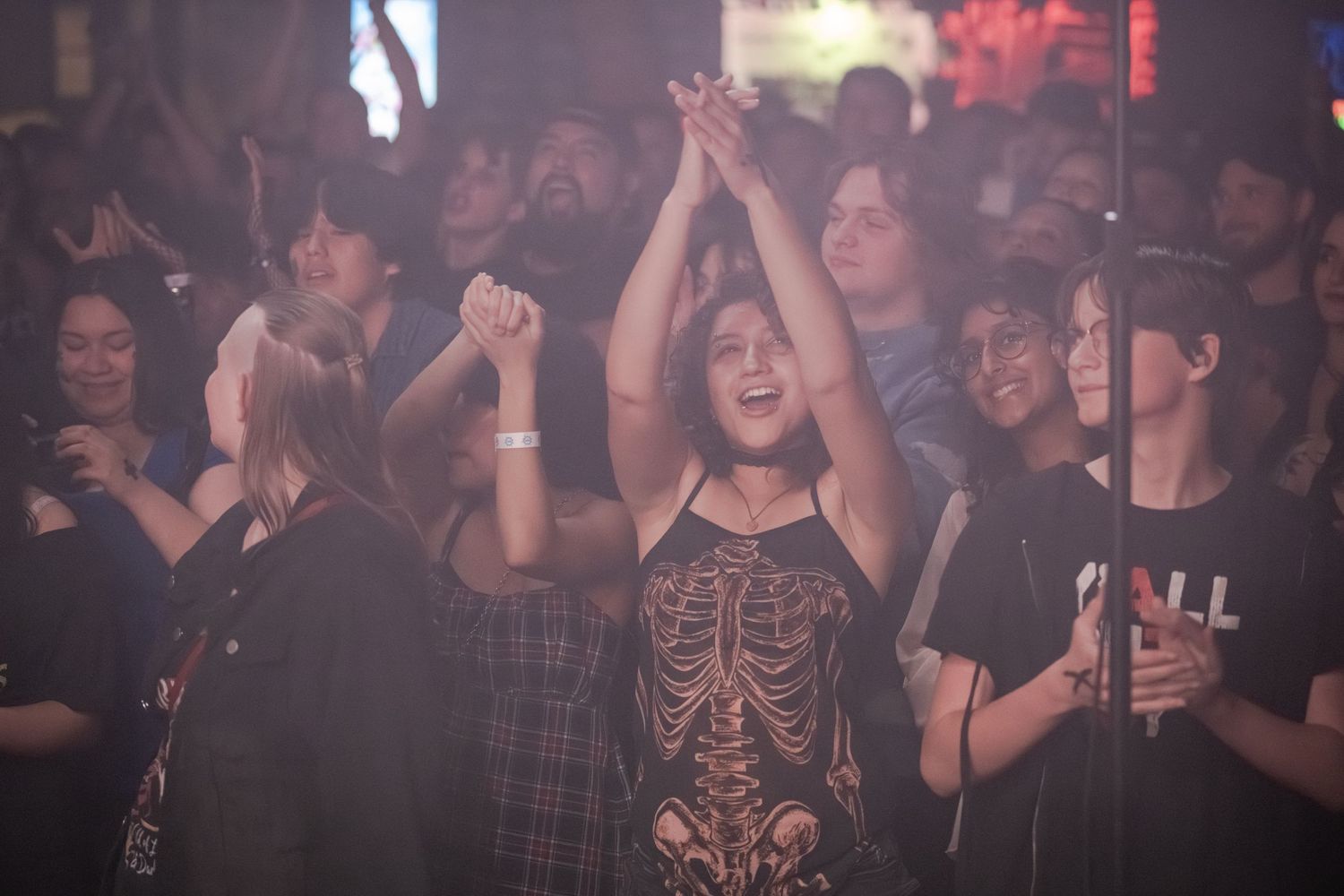 Excited crowd at a concert, woman in skeleton shirt raises hands joyfully amid energetic nightclub atmosphere.