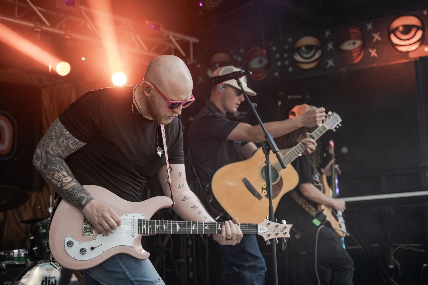 Rock band performing live on stage, guitarist with tattoos playing electric guitar under red stage lights.