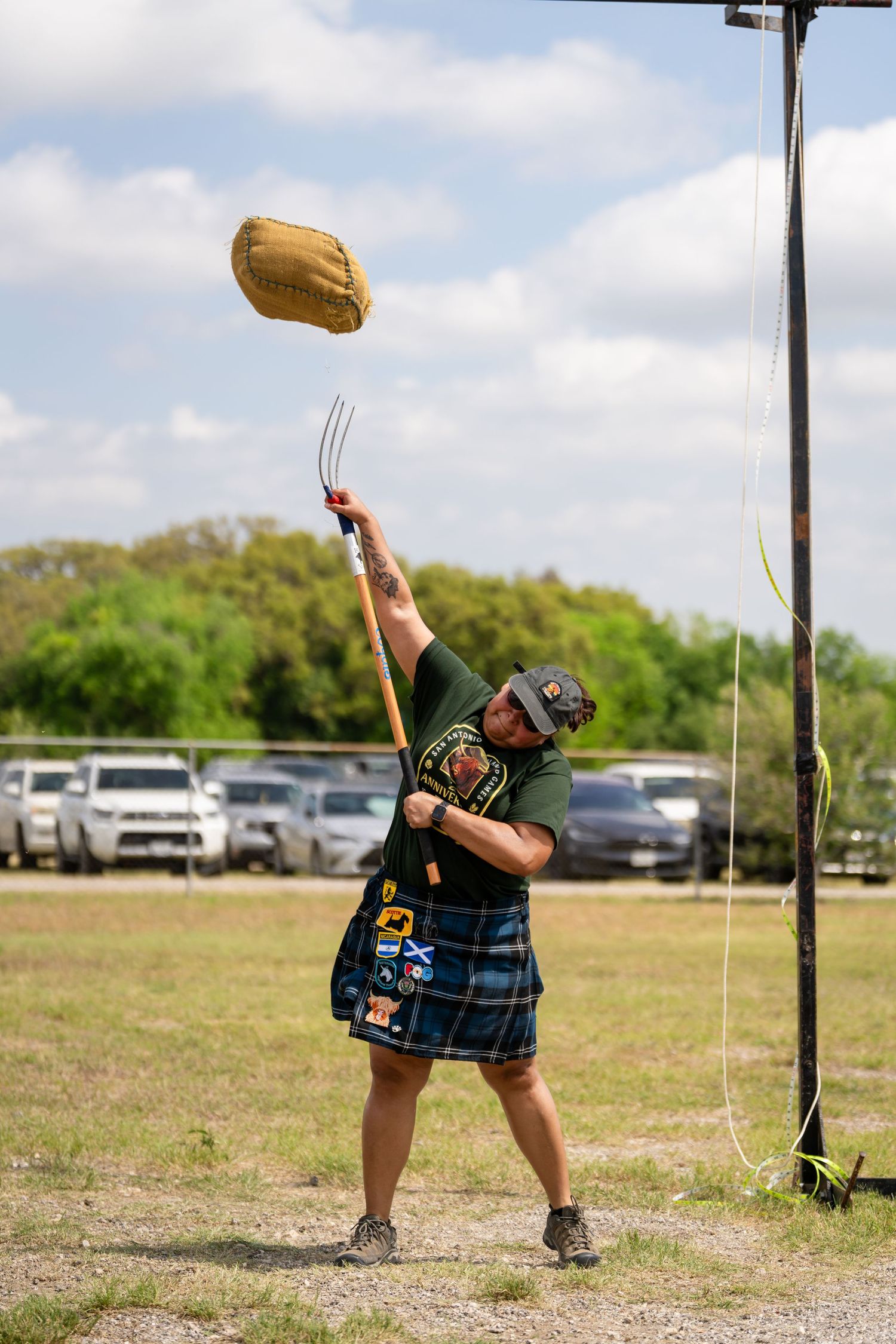 Man in kilt tossing sheaf with pitchfork at Highland Games competition outdoors.