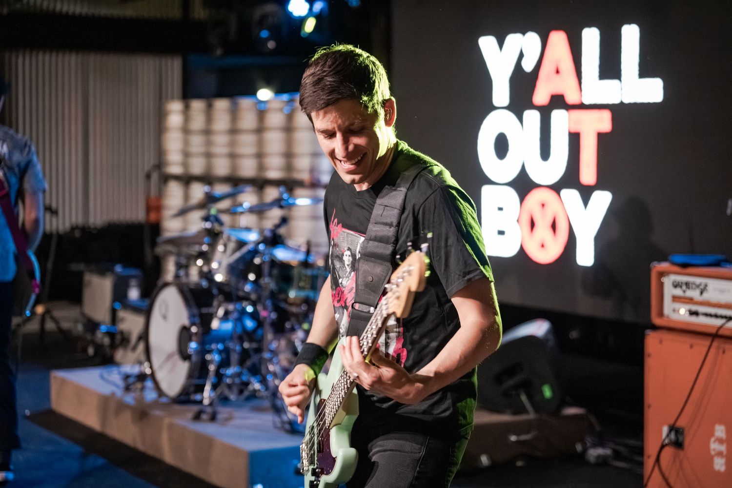 Guitarist performing live on stage in front of a Y'ALL OUT BOY neon sign at an indoor music venue.
