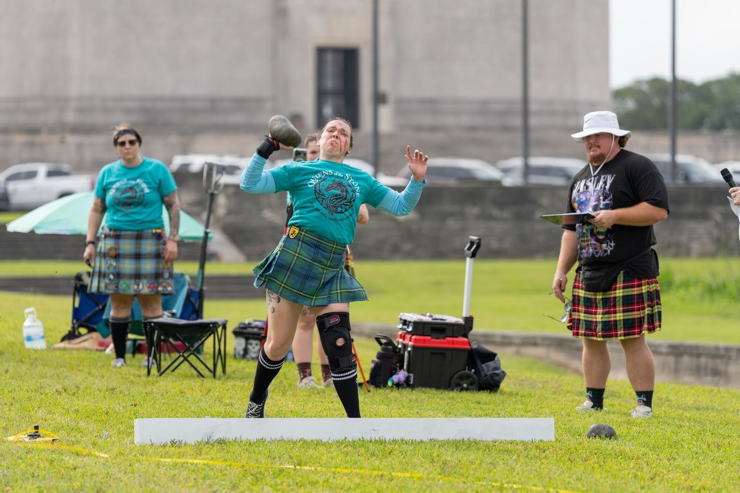 Woman competing in Highland Games shot put, wearing tartan kilt, with officials and spectators on green field.