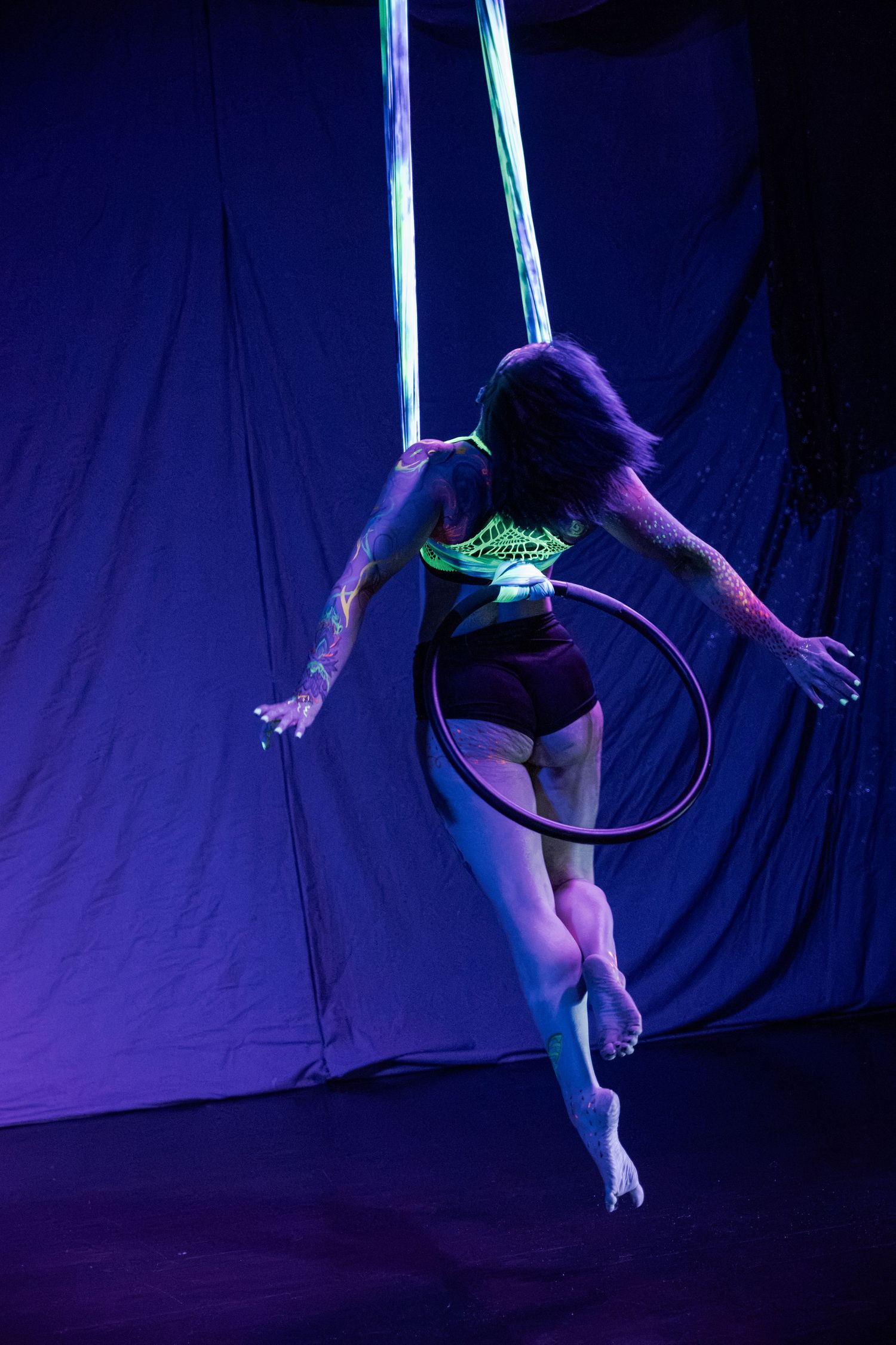 Aerial silk performer balancing a hula hoop under UV neon lights on a dark stage.