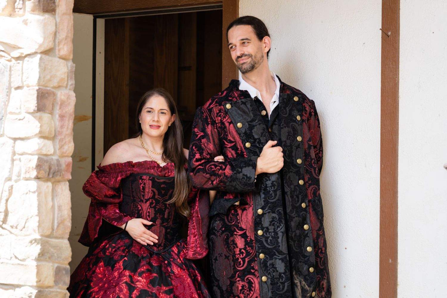 Couple in elegant Victorian-era red and black costumes posing in a doorway.