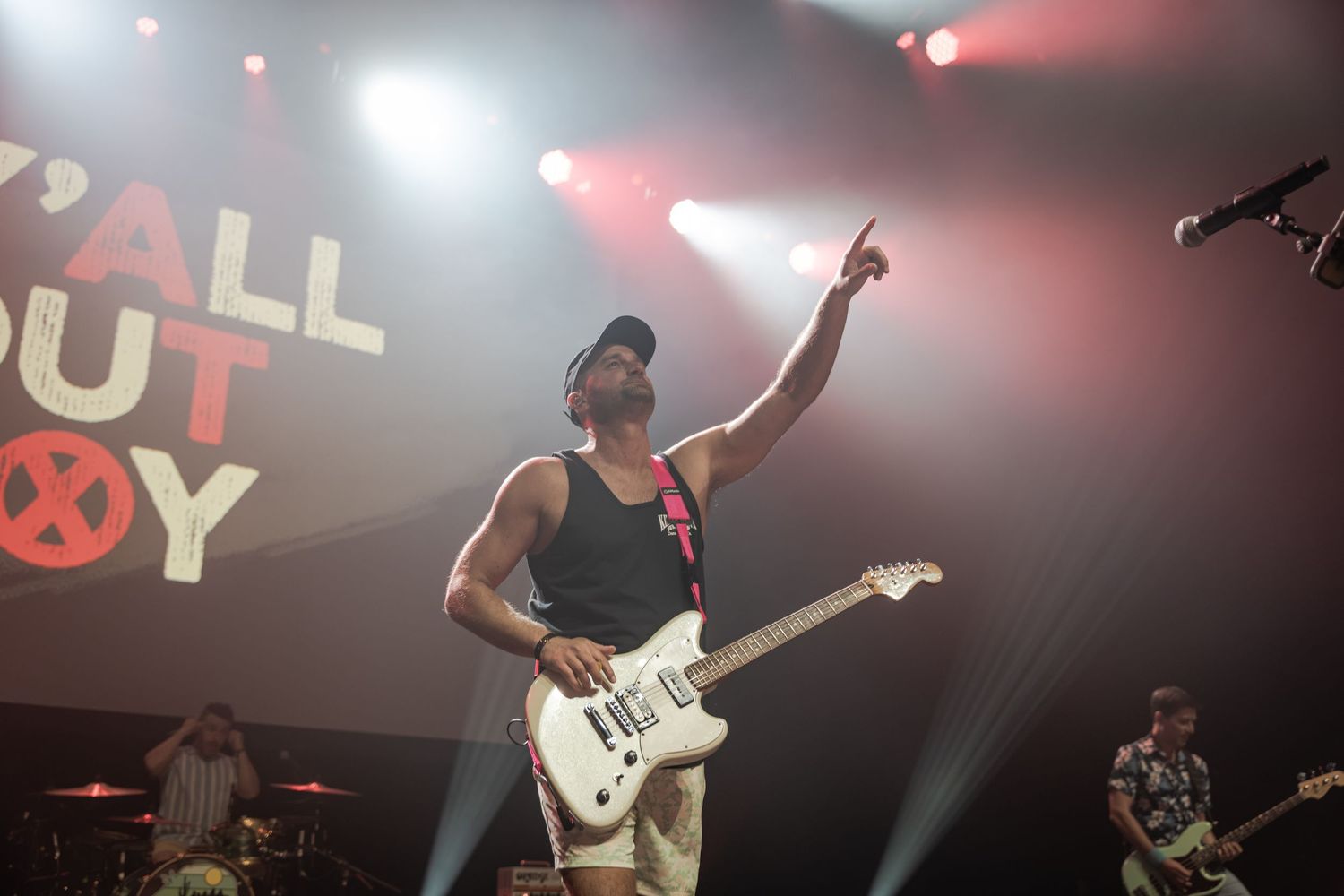 Fall Out Boy guitarist performs on stage, pointing upward under colorful stage lights with band logo displayed behind.