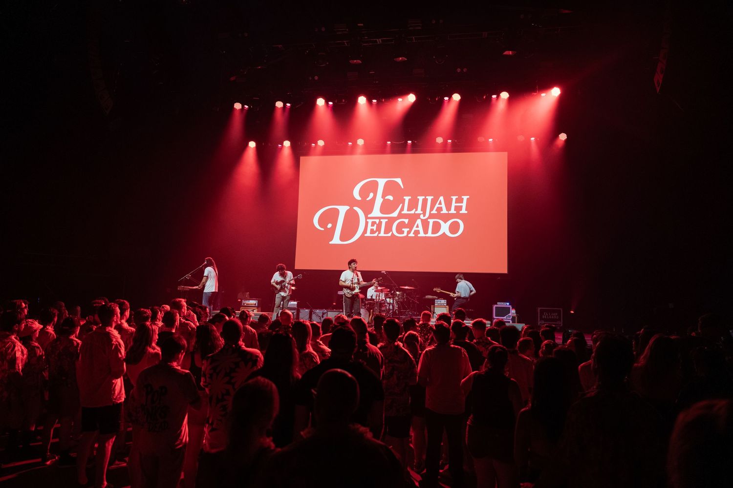 Elijah Delgado performing live on stage with red lighting and a large LED screen displaying his name to a standing crowd.