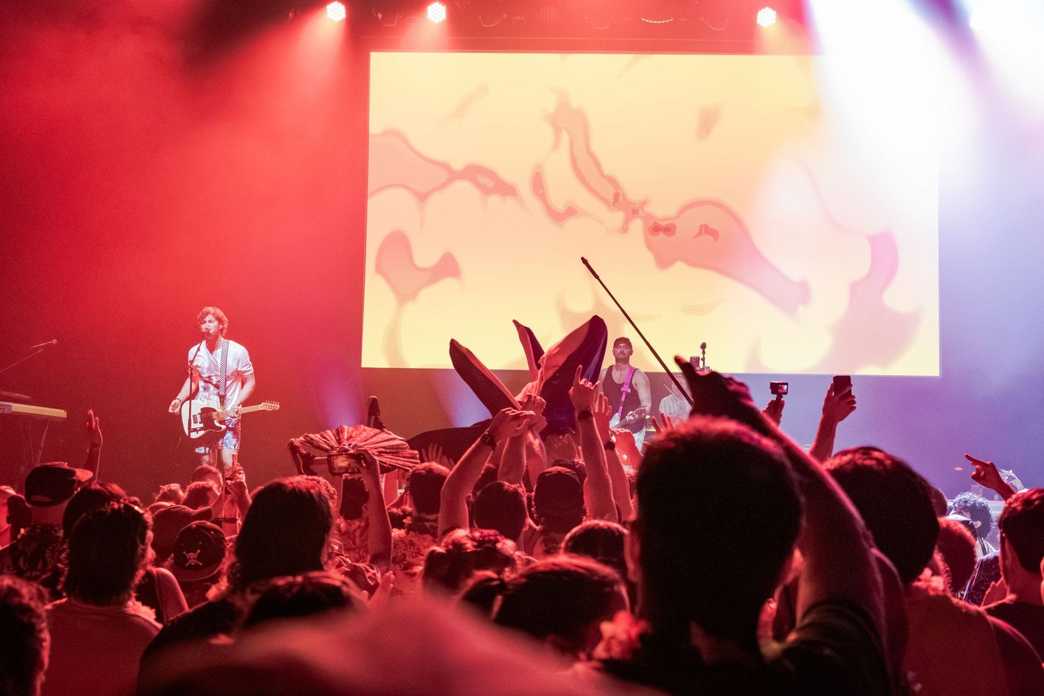 Live concert with band on stage, colorful projection screen, crowd cheering under red and pink stage lights.