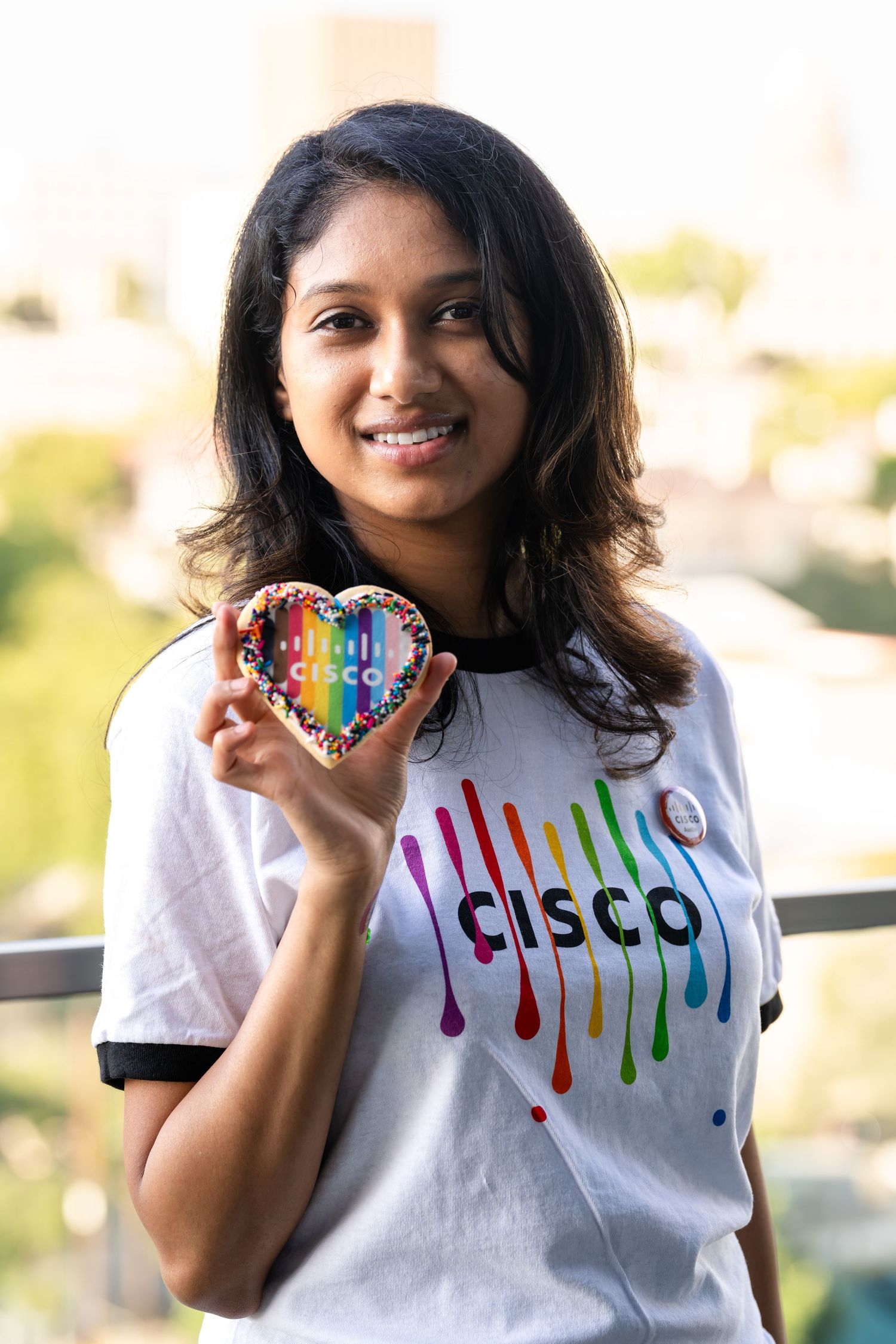 A smiling woman in a Cisco rainbow Pride t-shirt holds a heart-shaped rainbow Cisco cookie outdoors.