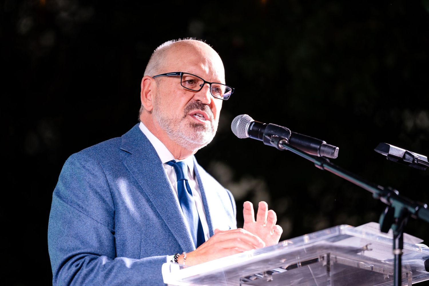 Speaker in blue suit and glasses gestures while speaking at podium with microphones during outdoor event.