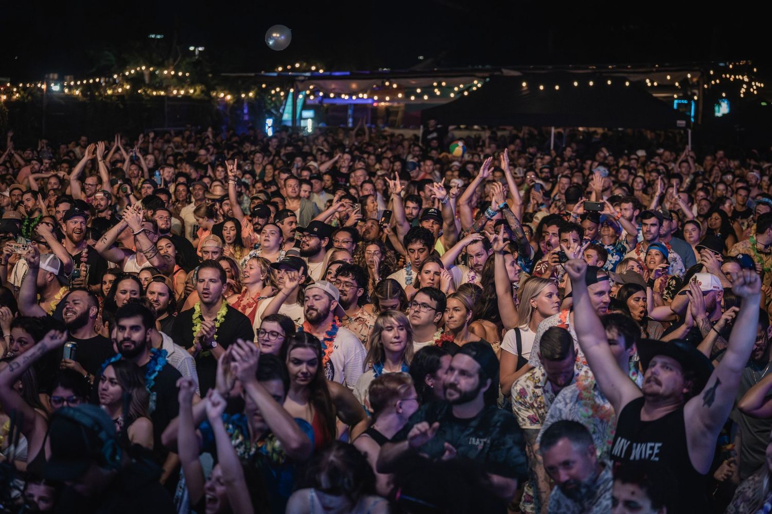 Energetic concert crowd with raised hands enjoying a live music performance in a dark venue with colorful lighting.