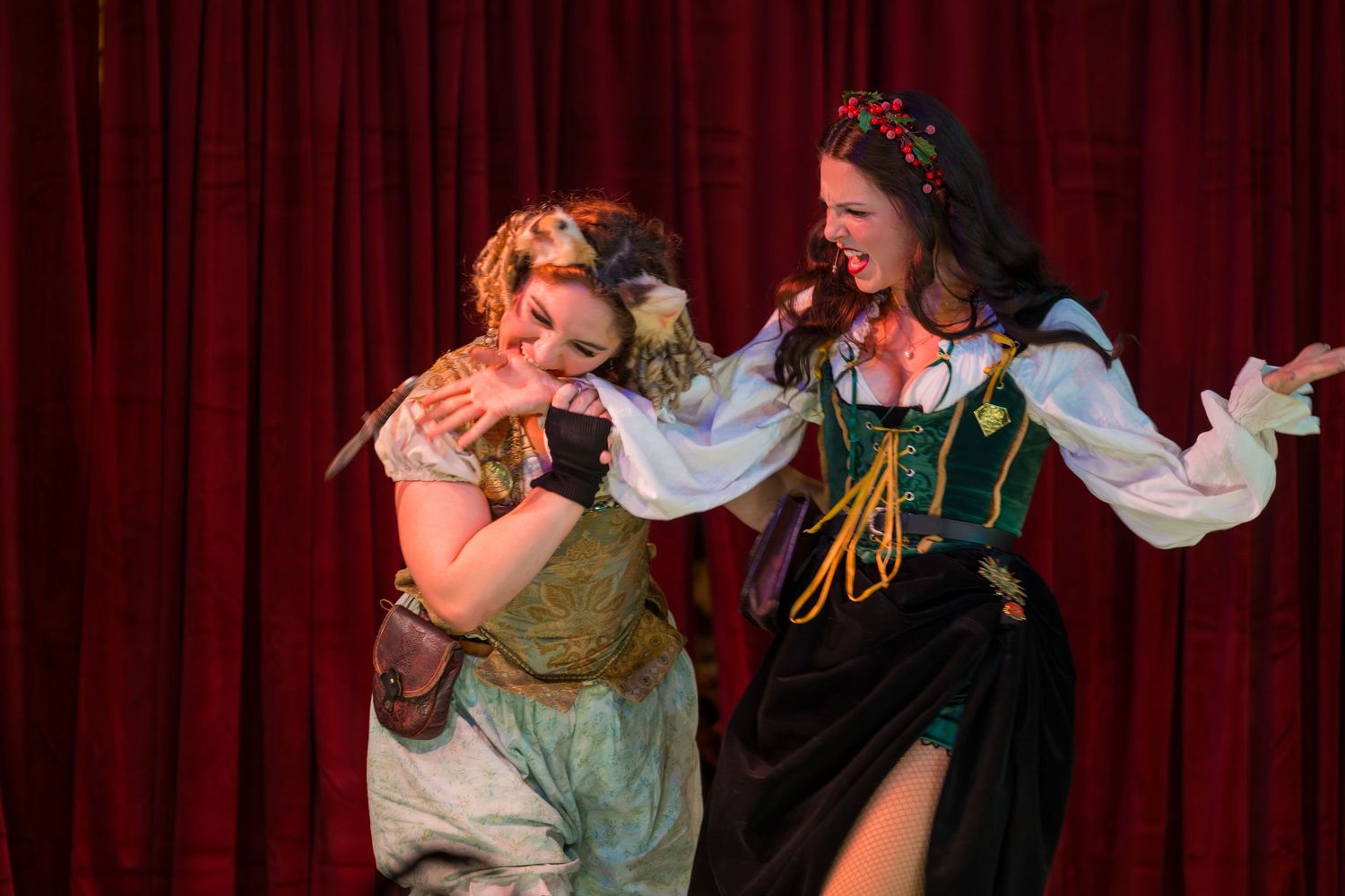 Two performers in medieval costumes act dramatically on stage with a red curtain backdrop during a theatrical production.