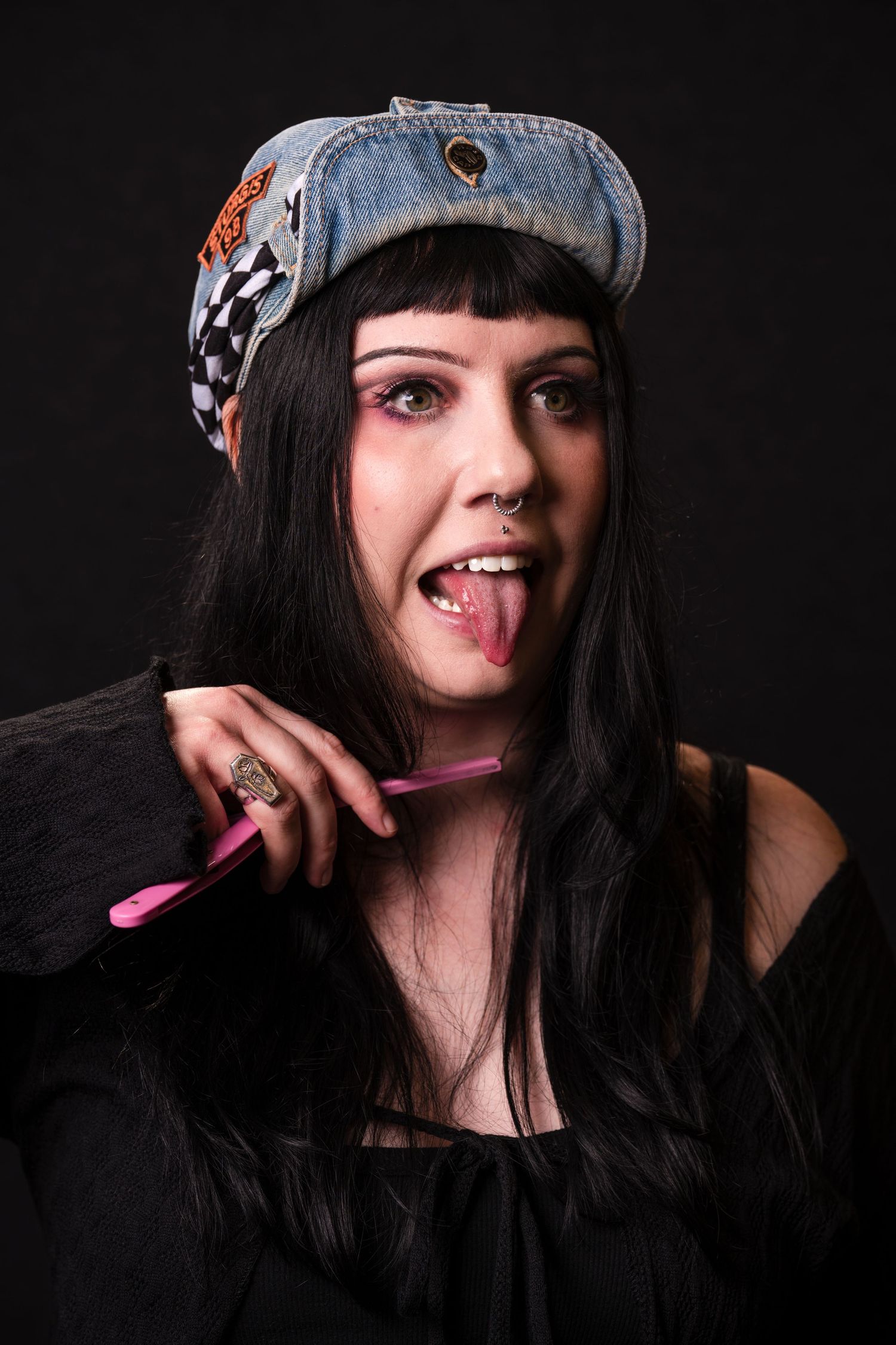 Person wearing a denim cap with patches sticks out tongue while holding a pink pen against dark background.