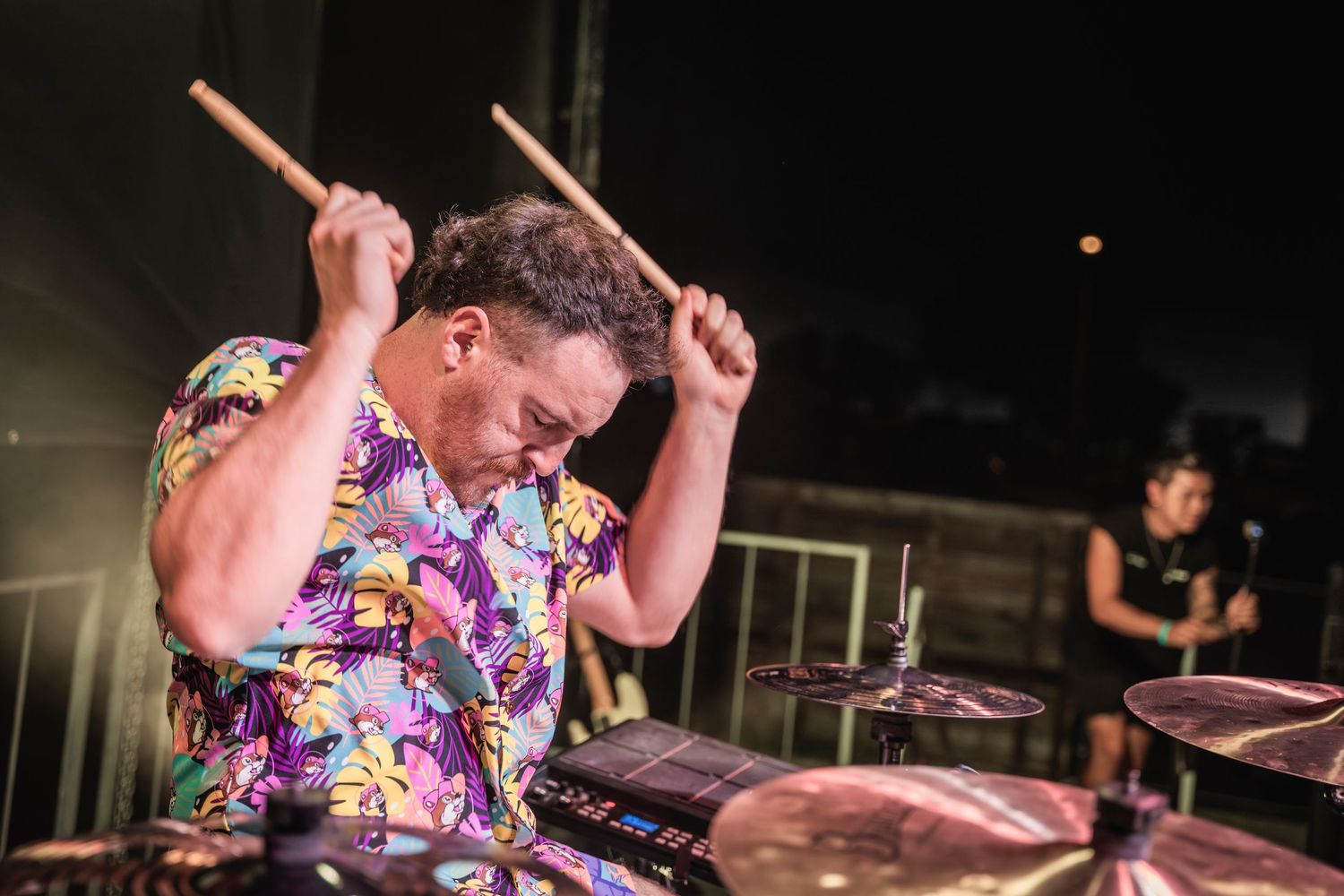 Drummer in colorful floral shirt performs energetically on stage during live music performance.