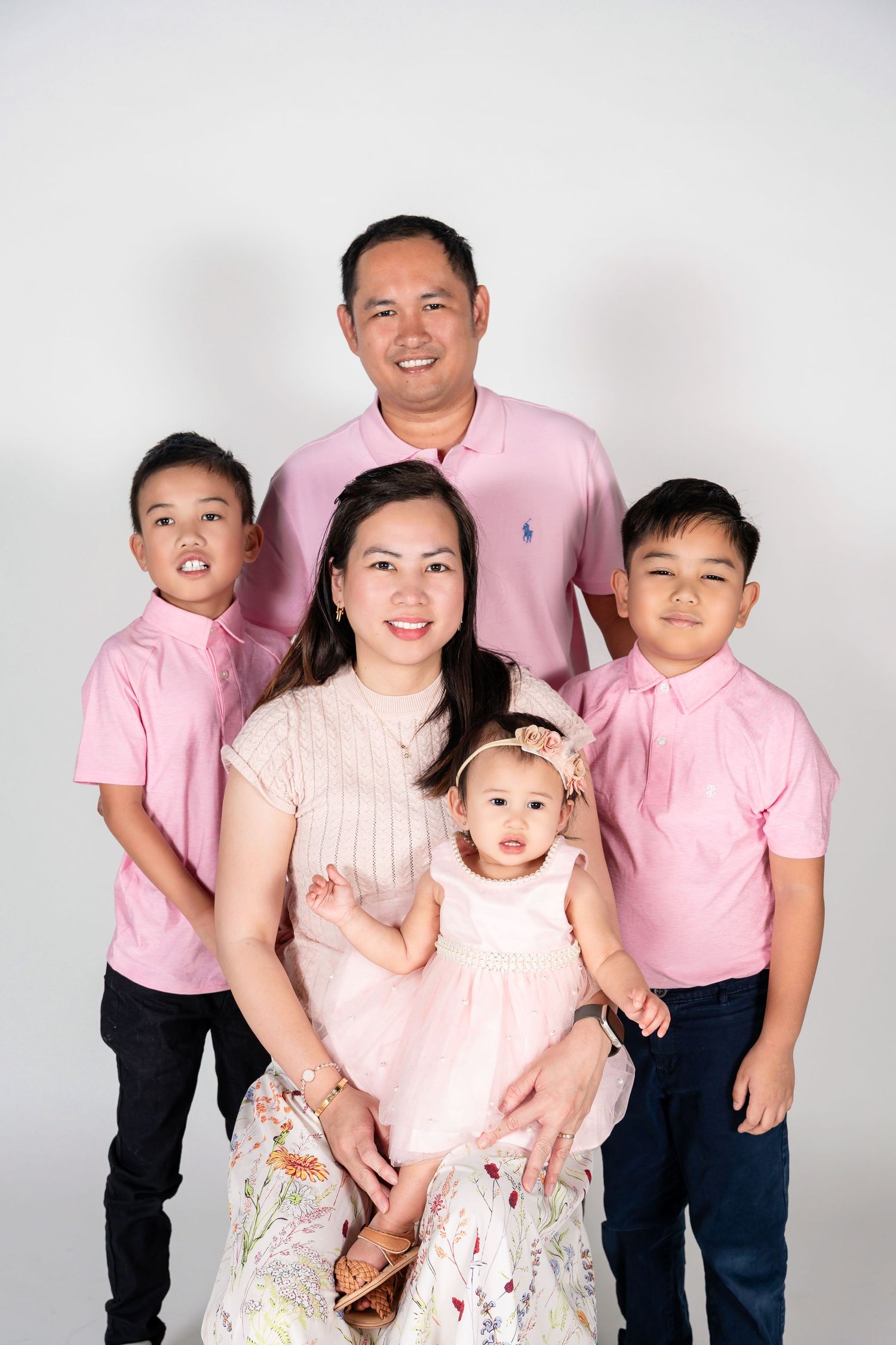 Asian family of five in matching pink outfits posing for a studio portrait, parents with two boys and a baby girl.