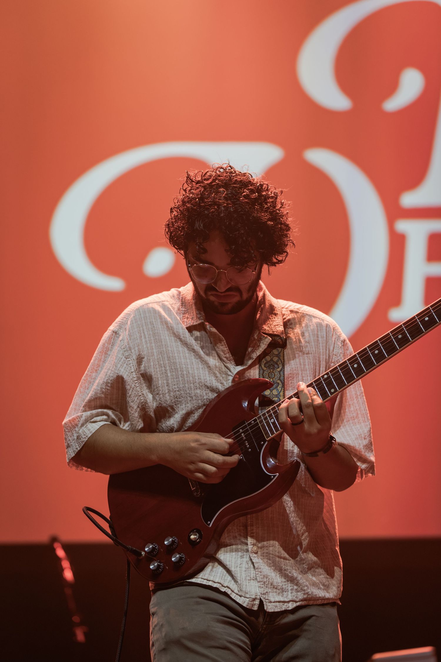 Curly-haired guitarist plays electric guitar on stage with red-lit backdrop during live music performance.