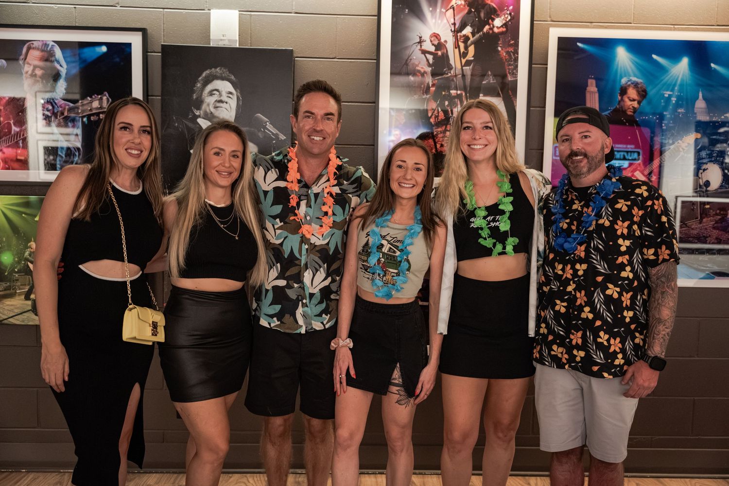 Group of six friends posing together at a music venue with concert photos on the wall behind them.