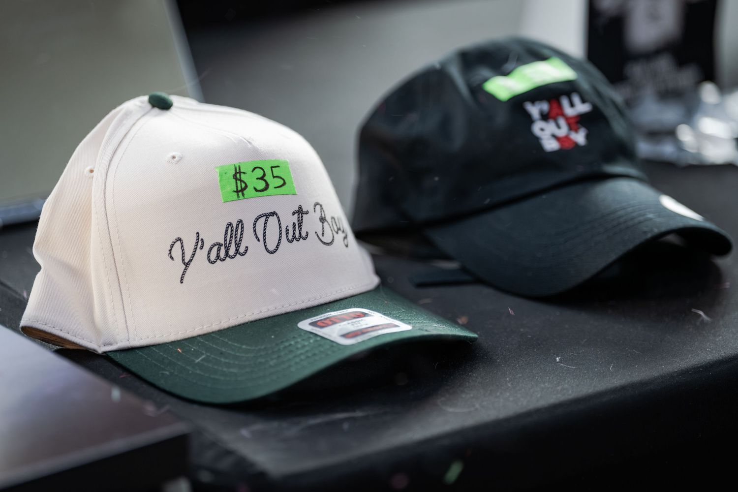 Two baseball caps on display, one white with green brim reading Y'all Out Boy priced at $35, and one black cap.