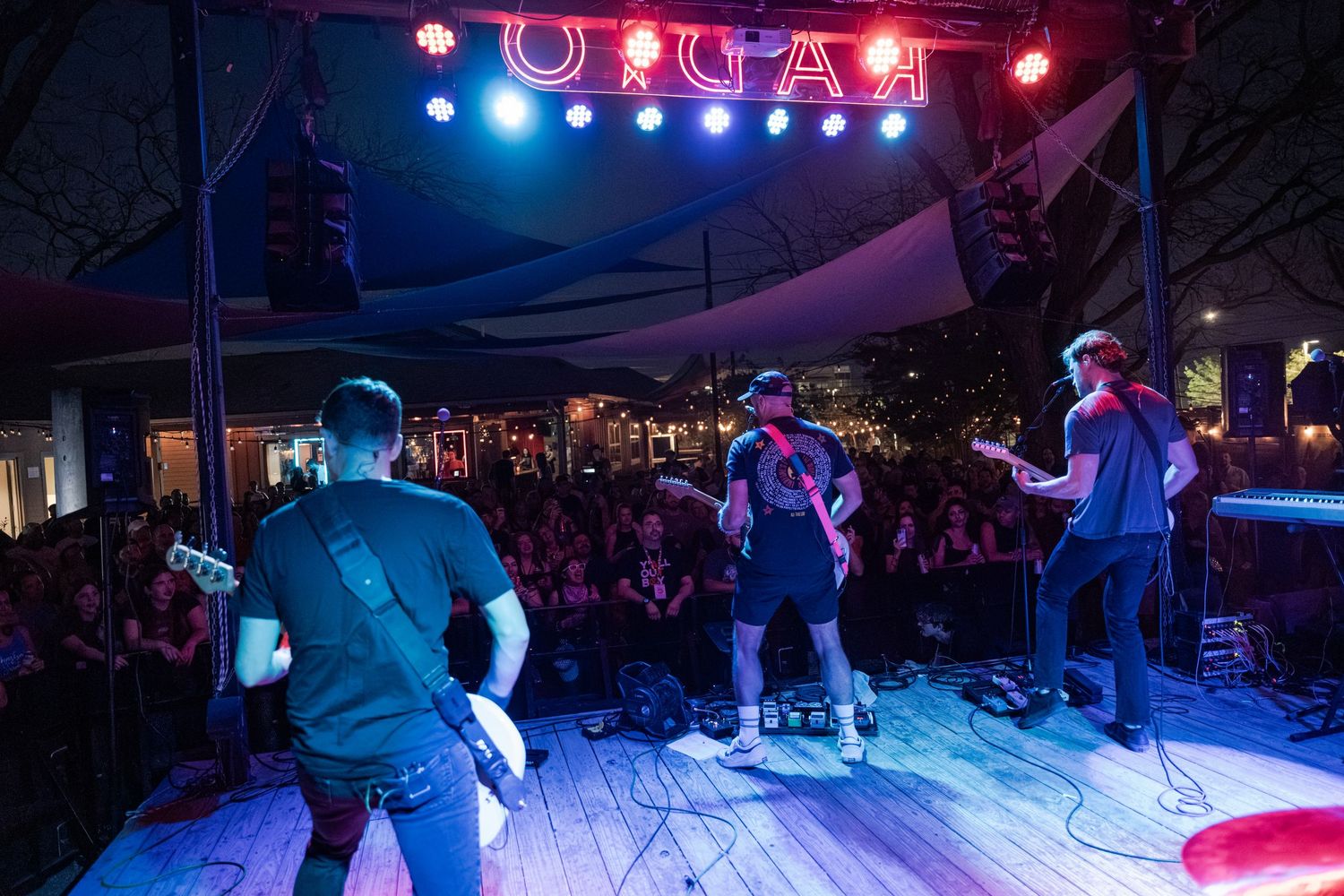 Three musicians perform on an outdoor stage at night before a large crowd under colorful lights at ORGAA.