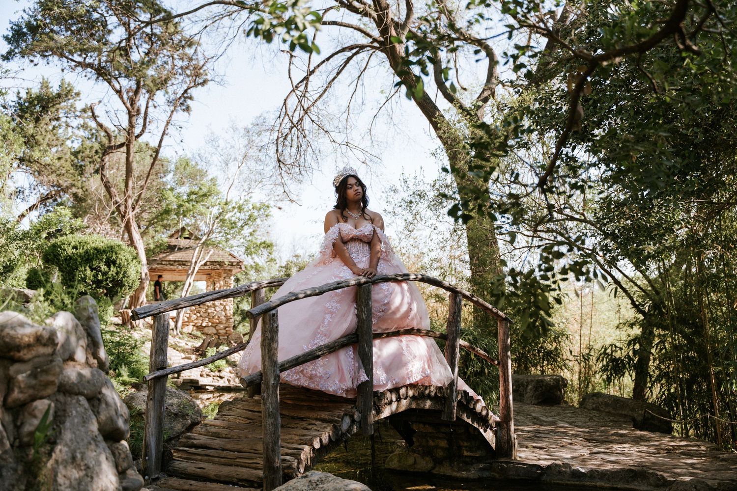 Young woman in a pink ball gown poses gracefully on a rustic wooden bridge surrounded by lush green trees.