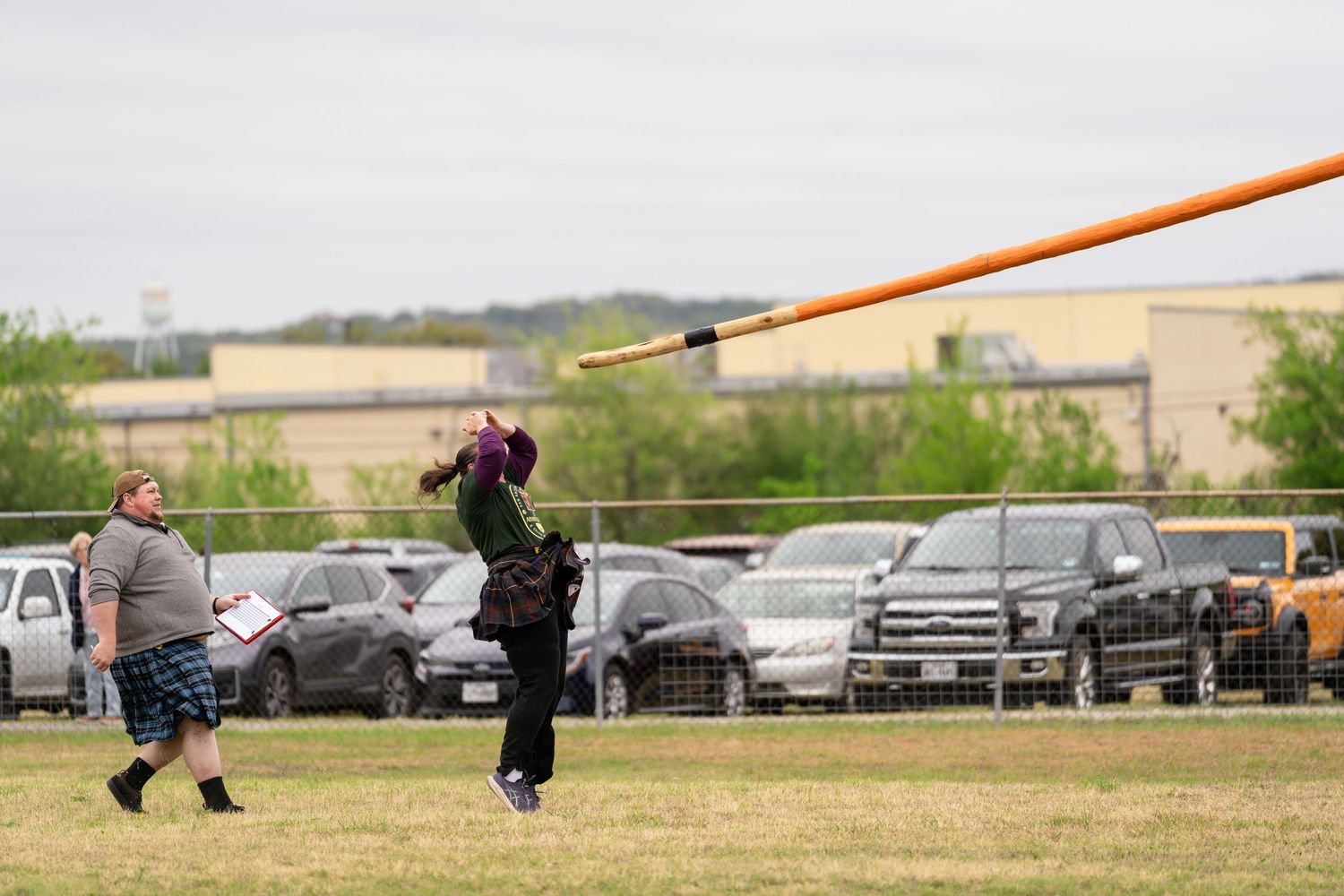 Athletes compete in caber toss at a Highland Games event on a green field with cars parked in the background.