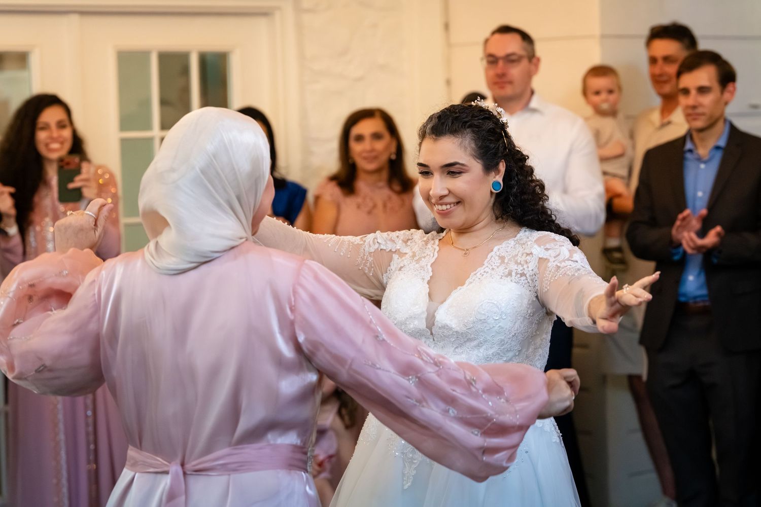 Bride in white lace gown dancing joyfully with a guest in pink hijab at a wedding reception.