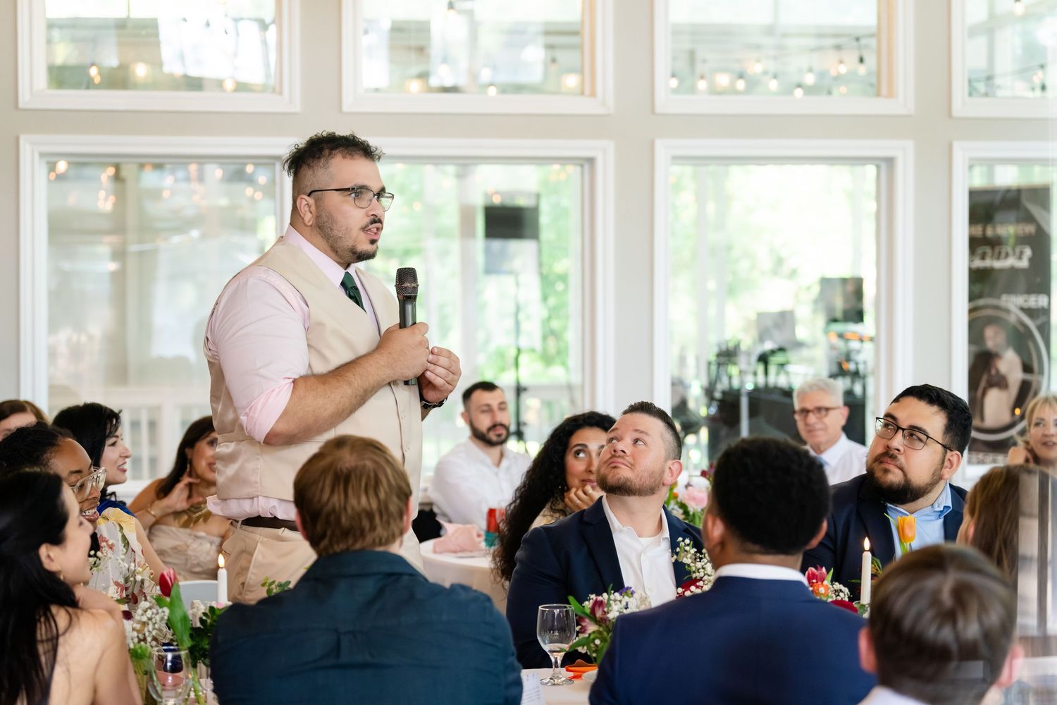 Man giving a speech with microphone at an elegant wedding reception while guests listen and smile at decorated tables.