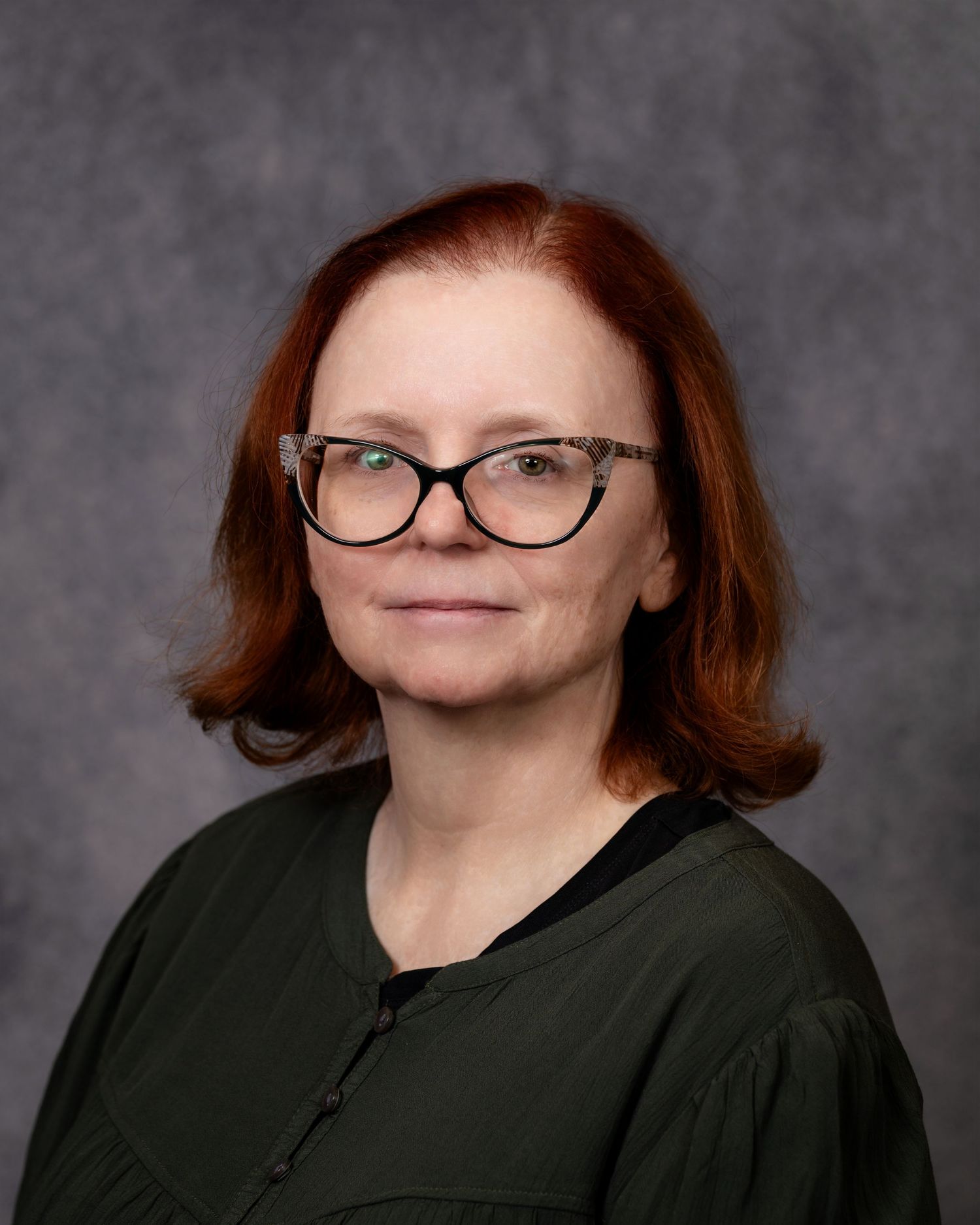 Professional headshot of a woman with red hair and glasses wearing a dark green top against a gray background.