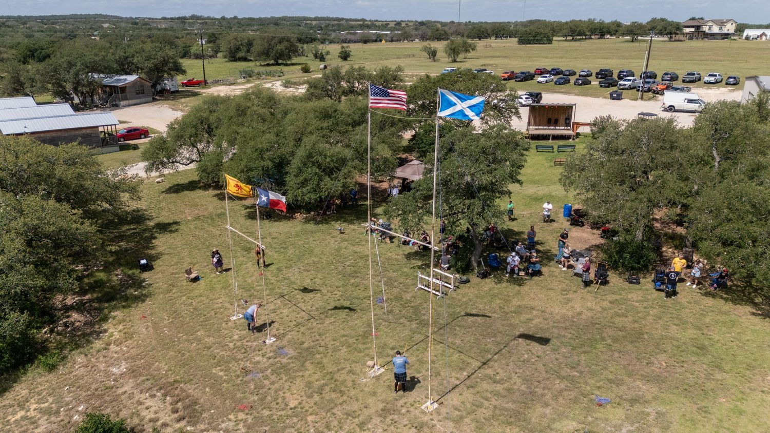 Aerial view of outdoor gathering with American and Scottish flags on tall poles in a green field with trees and parked cars.