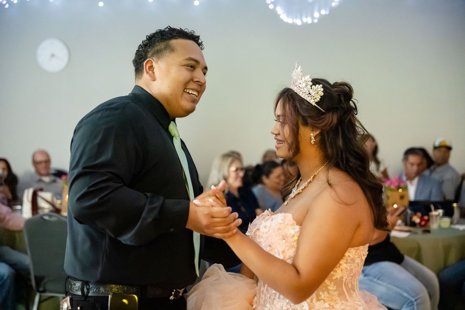 A young man and woman share a joyful dance at a quinceañera celebration, with the birthday girl wearing a tiara.