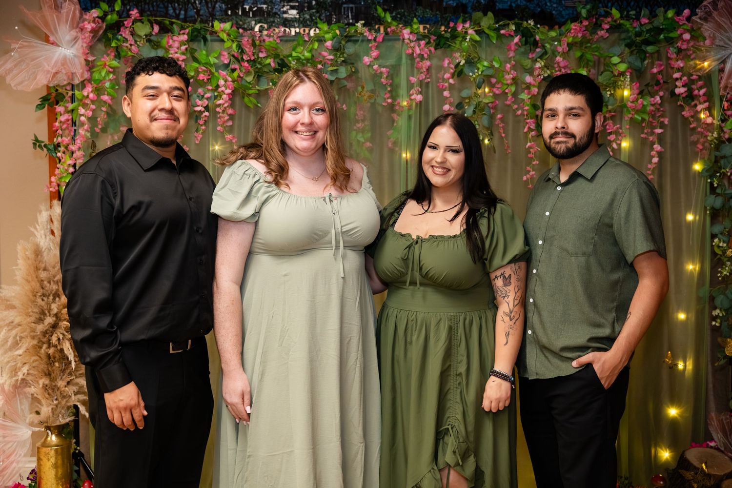 Four people smiling together in front of a floral decorated backdrop with fairy lights at a festive event.