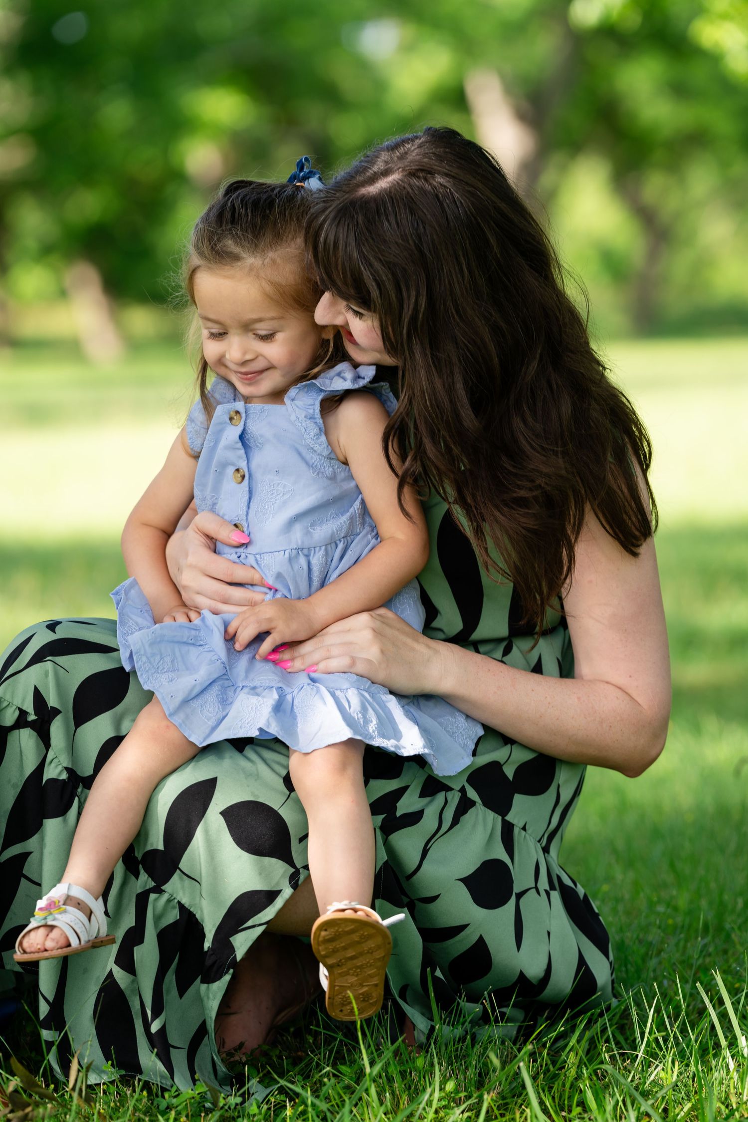 A mother in a green floral dress holds her smiling toddler girl in a blue dress outdoors in a lush green park.