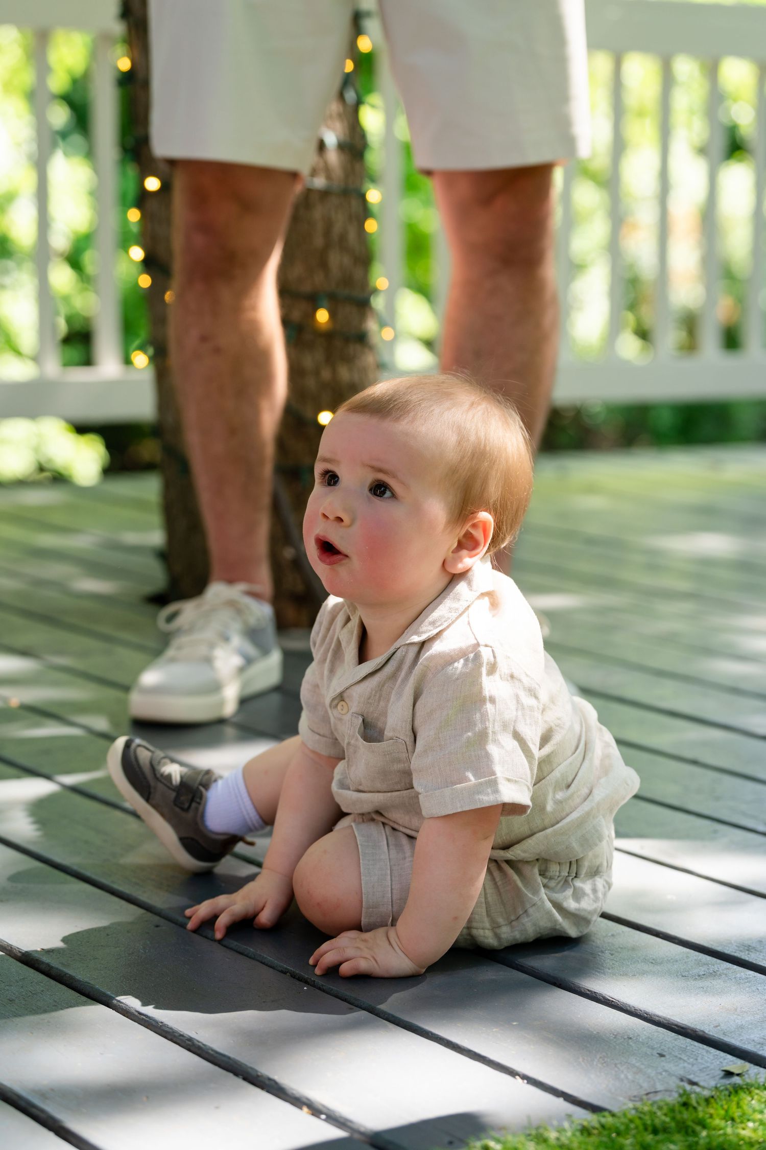 Adorable baby boy in beige romper sitting on outdoor deck, looking up curiously with adult standing nearby.