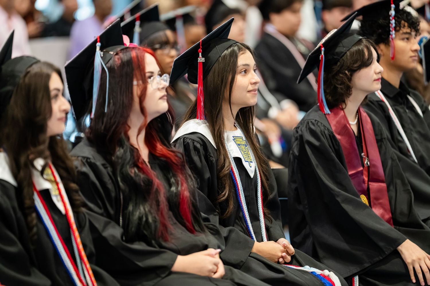 Students in graduation caps and gowns seated at a commencement ceremony, smiling and celebrating their achievement.