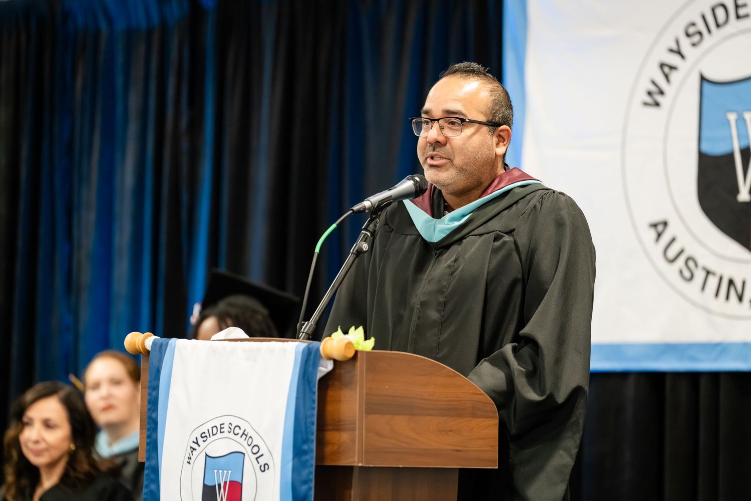Academic official in graduation regalia speaks at podium during Wayside Schools Austin commencement ceremony.