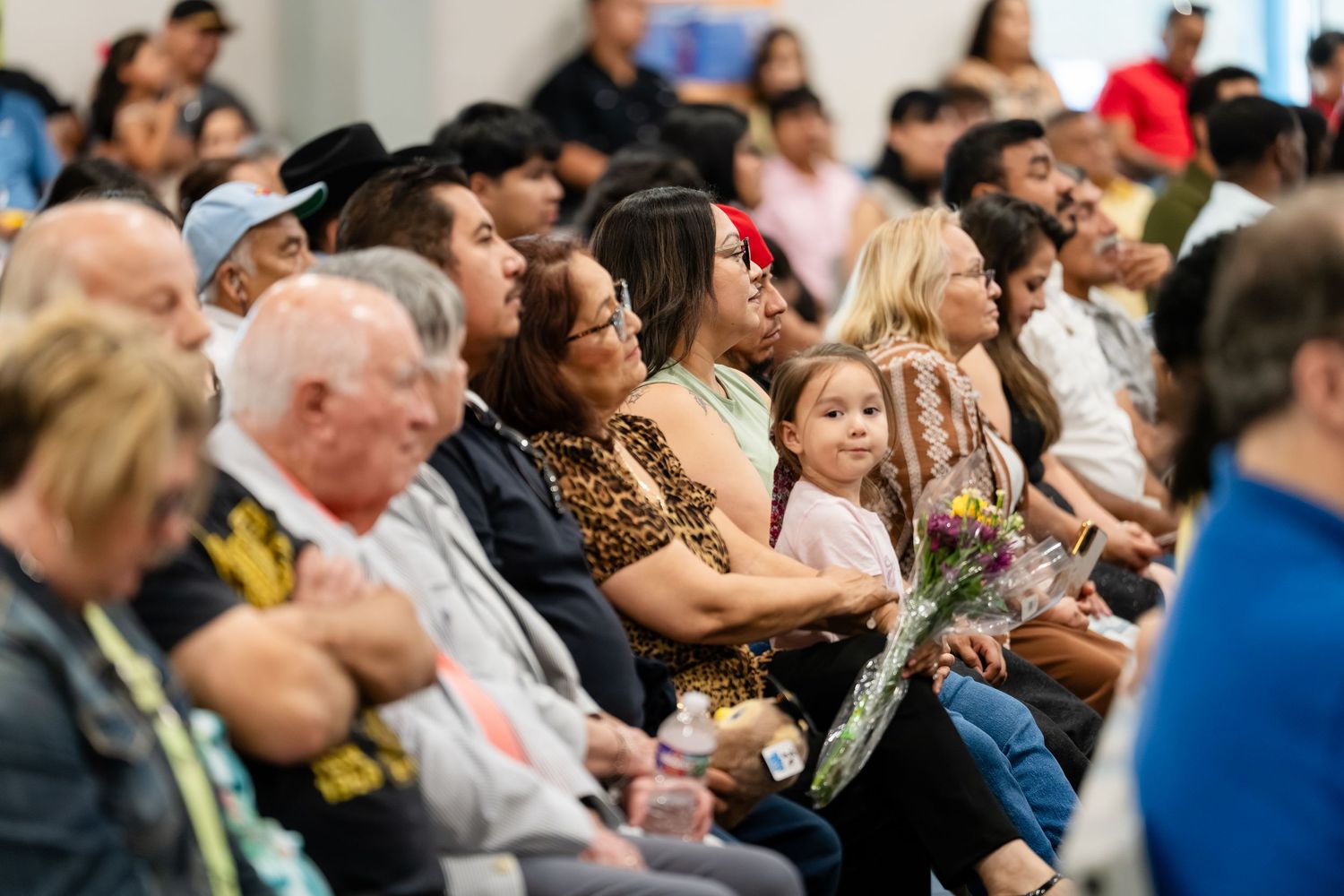 Diverse crowd of families seated together at an indoor community event, with a young girl holding yellow flowers.