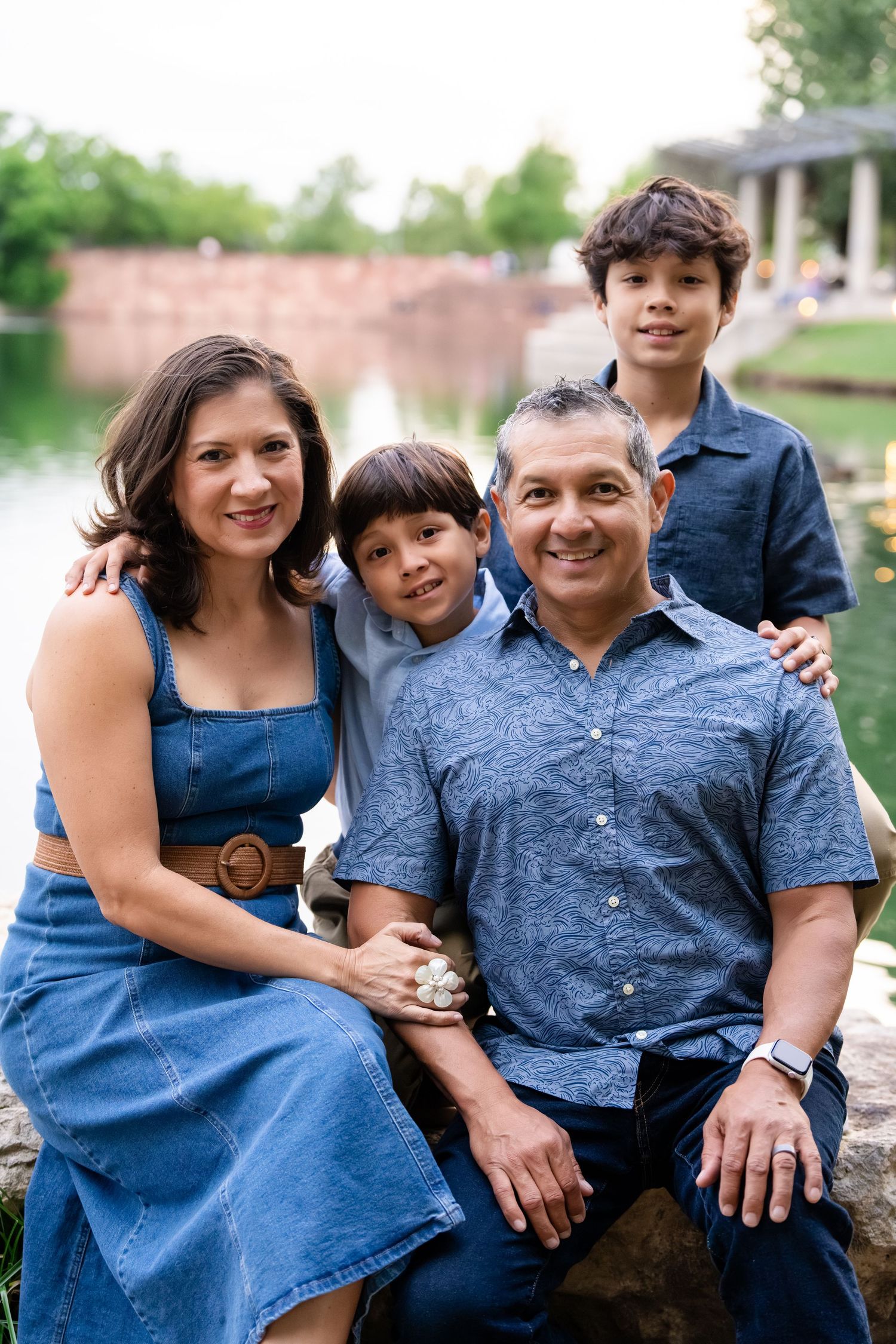 A smiling Hispanic family of four poses outdoors near a pond, wearing coordinating blue outfits for a portrait.