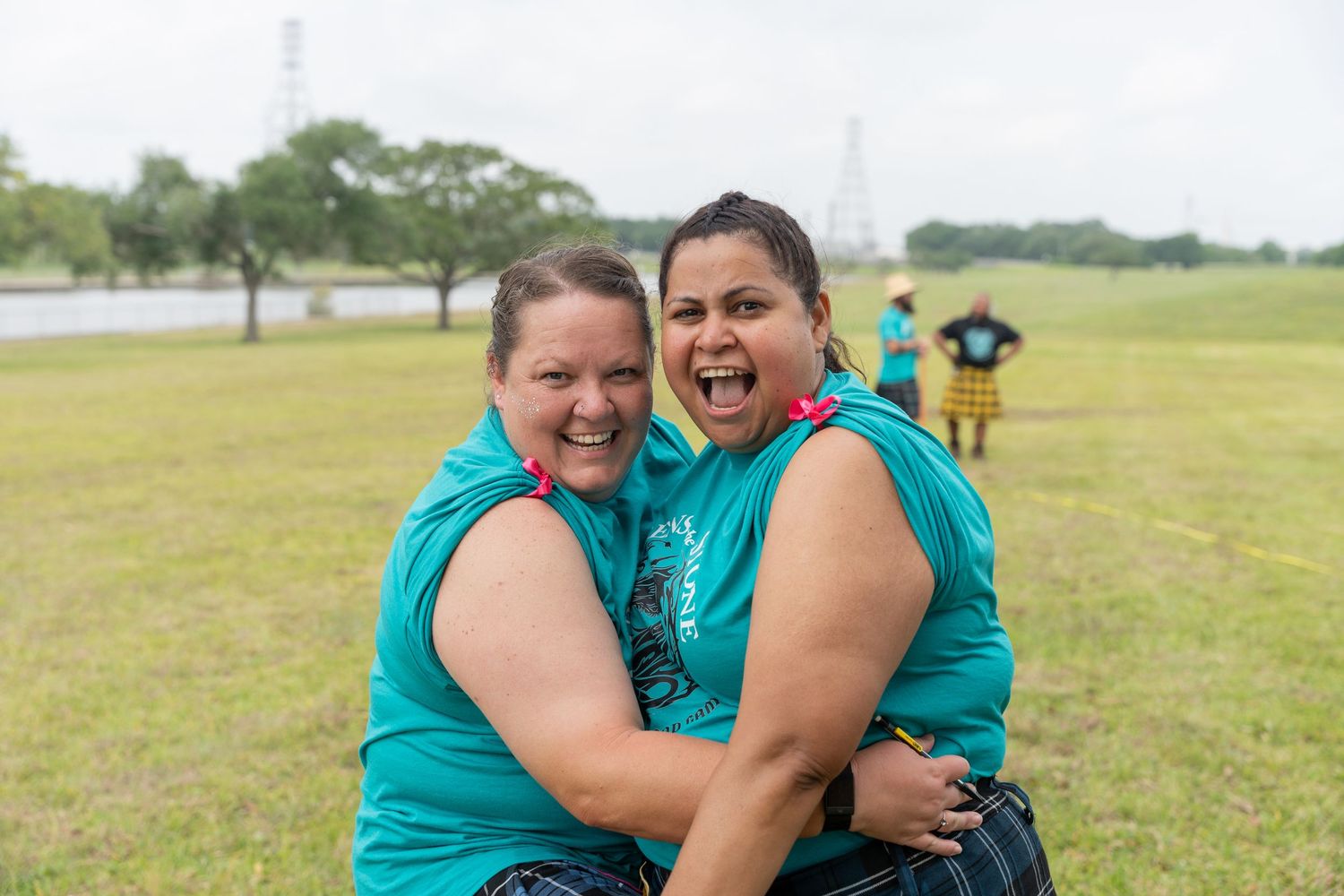 Two women in teal shirts laugh and hug outdoors on a green field during a fun event.
