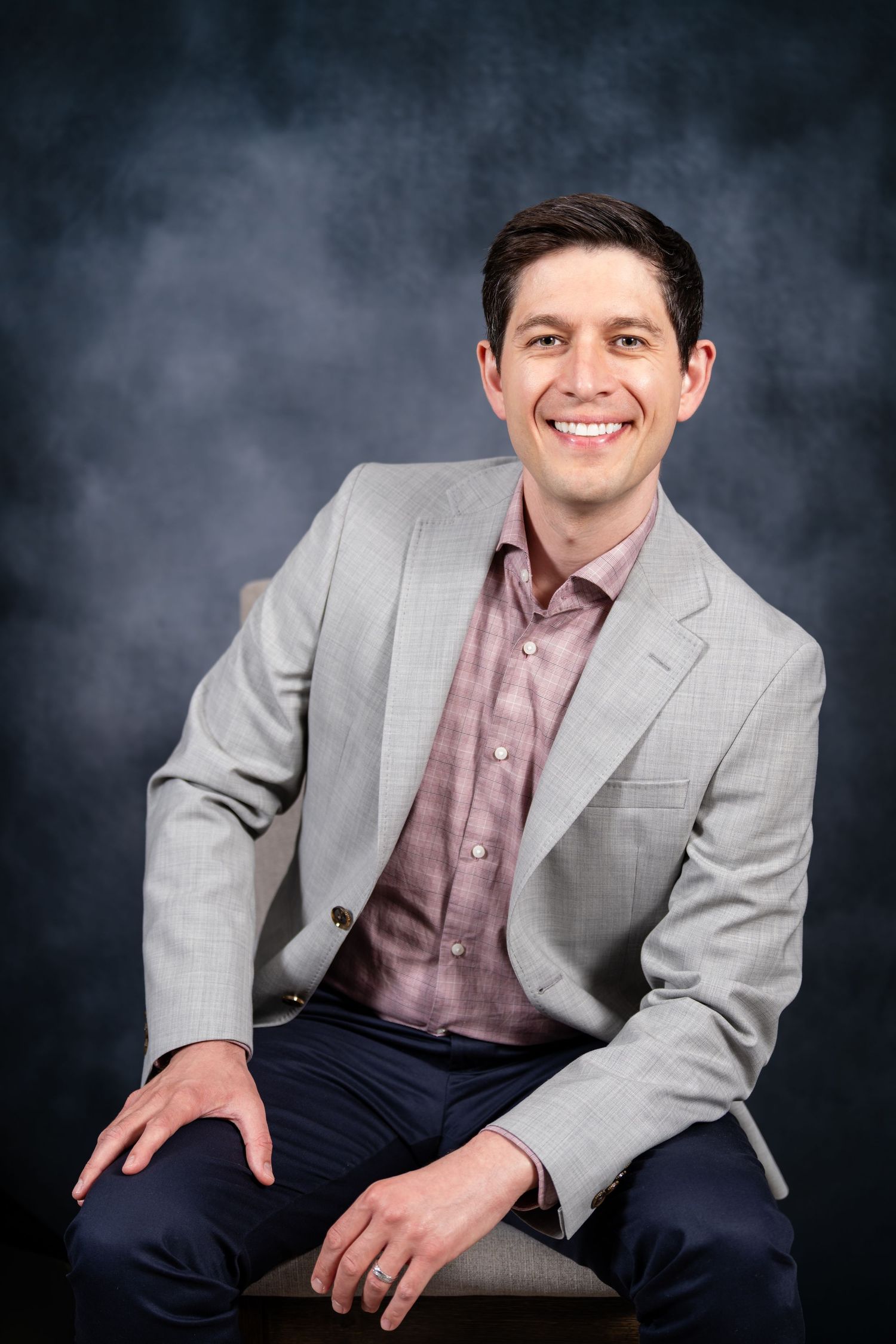 Smiling man in gray blazer and pink shirt seated confidently against a dark studio background.