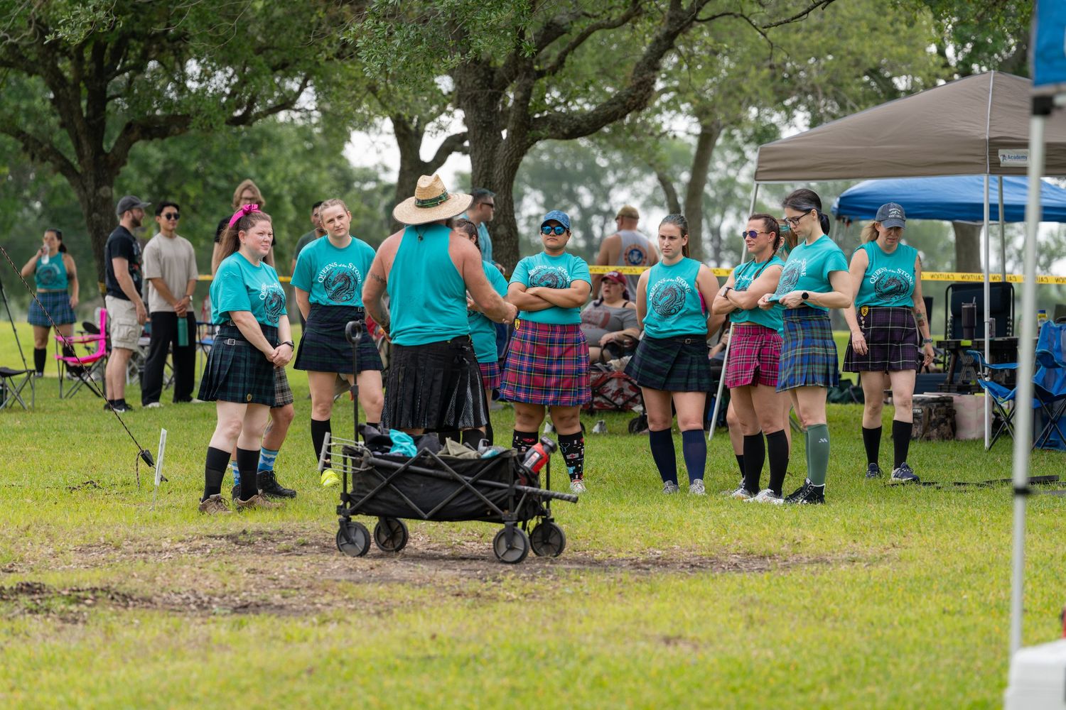 Group of people in teal shirts and kilts at a Scottish Highland Games event on a grassy field.