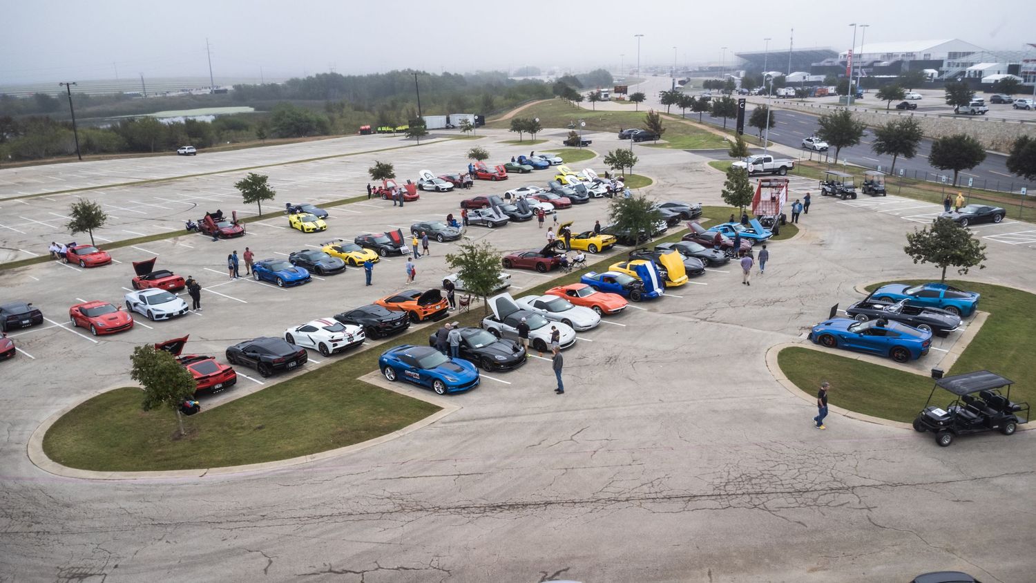 Aerial view of a car meet showing colorful vehicles arranged in curved rows in a parking lot.