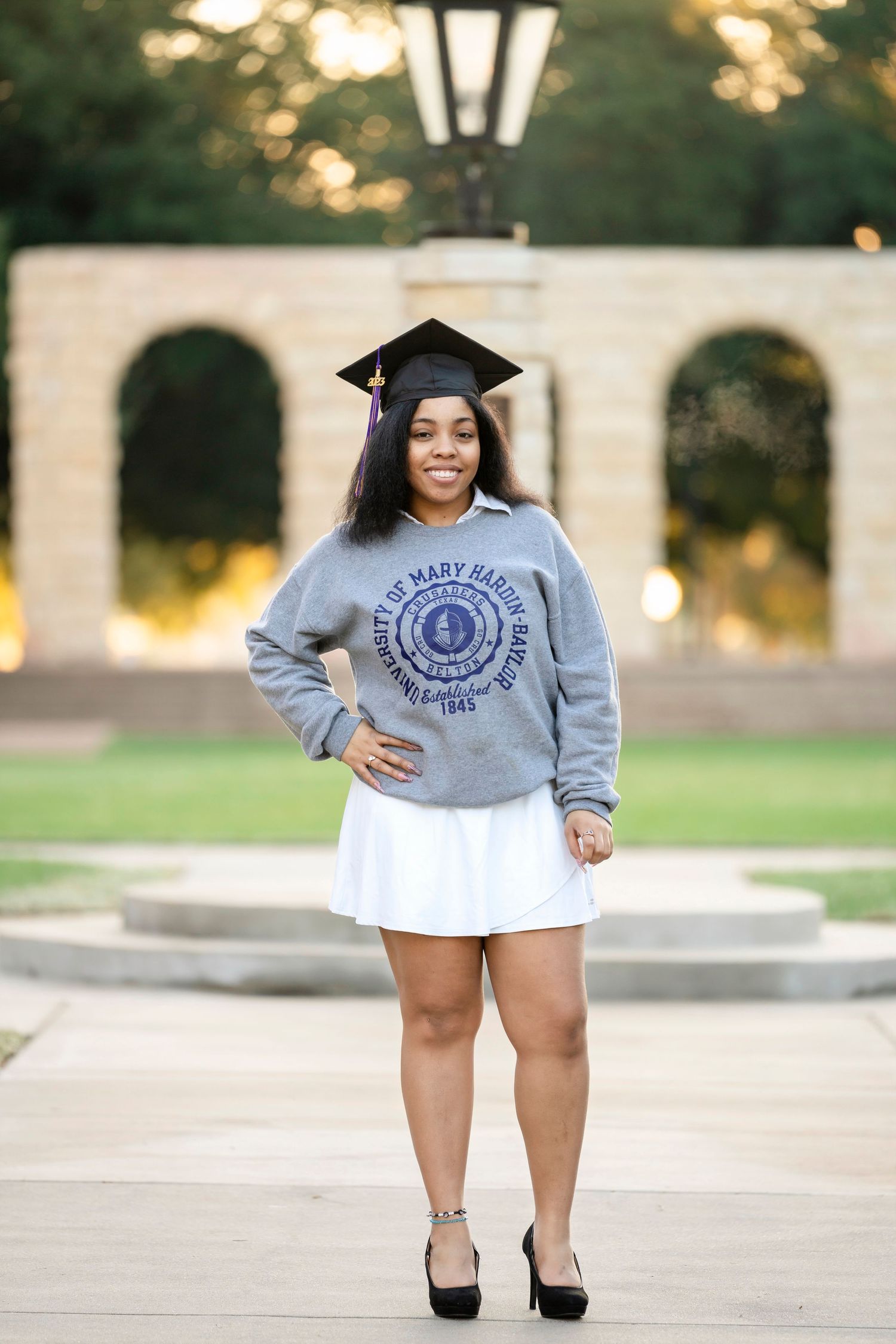 Graduate in cap and gown poses in front of campus archway at sunset.