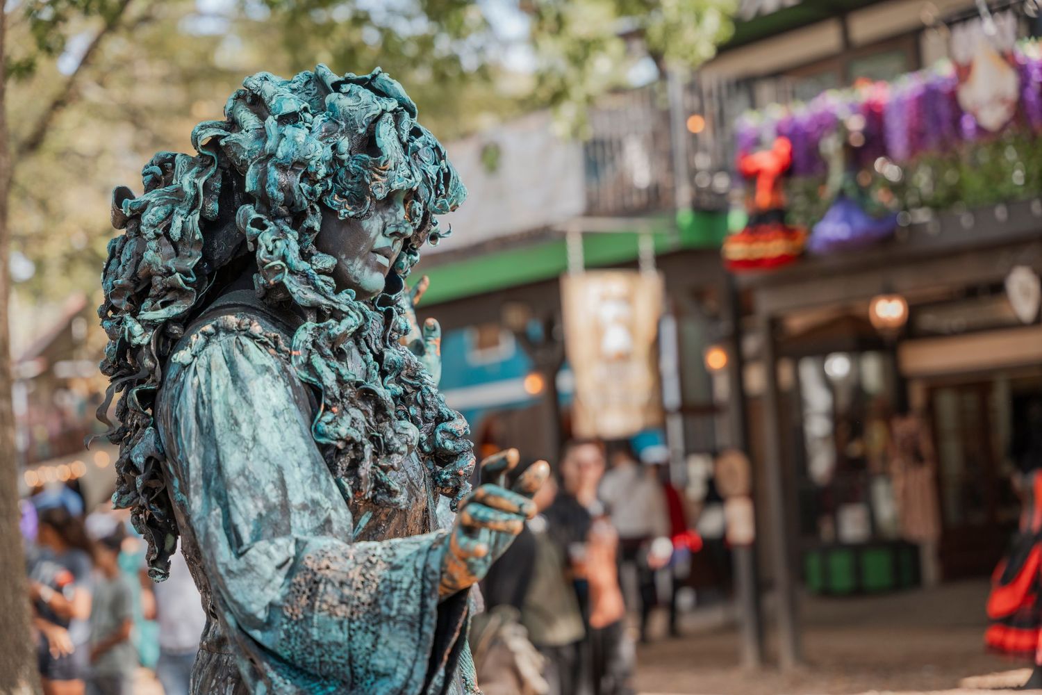 Weathered green patina statue at renaissance festival with shopping stalls in background.