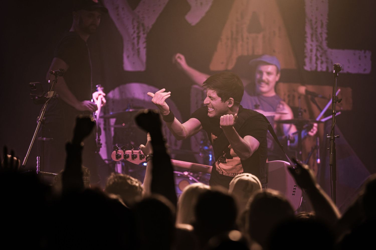 Musicians performing energetically on stage with crowd silhouettes visible in the foreground.