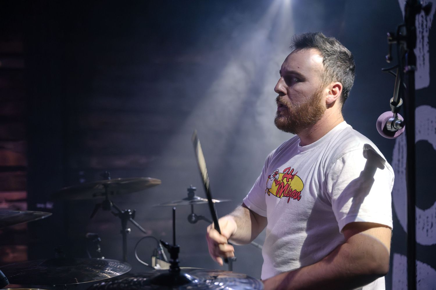 A drummer in a white t-shirt performs passionately on stage with dramatic lighting effects.