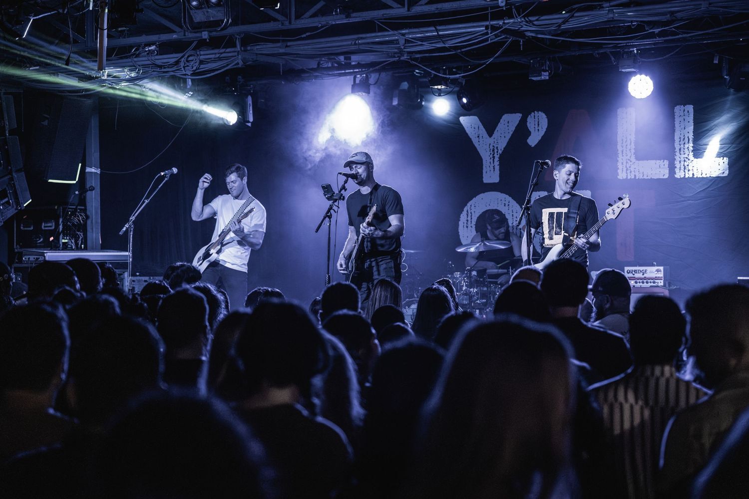 Rock band performing on concert stage with purple lighting and silhouetted audience below.